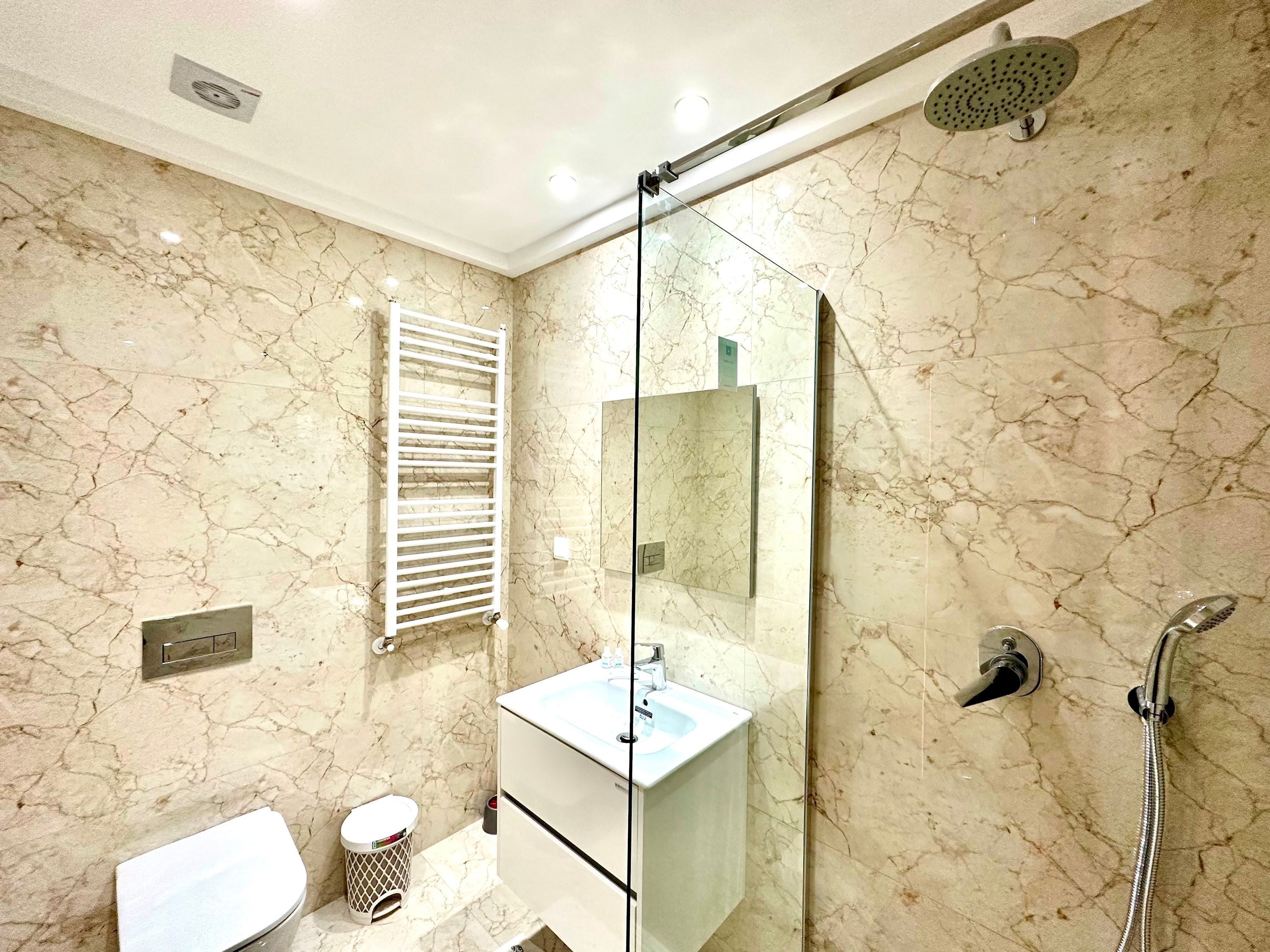 Een badkamer met een marmeren wand, een wit wastafelmeubel, een spiegel, een douche en een toilet.
