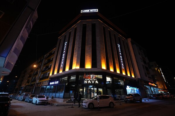 A hotel with a prominent sign reading "The NAVA Hotel" and "NAVA Cafe" on the ground floor. There are cars parked in front, and the building is illuminated with warm lighting. - THE NAVA HOTEL (Van)
