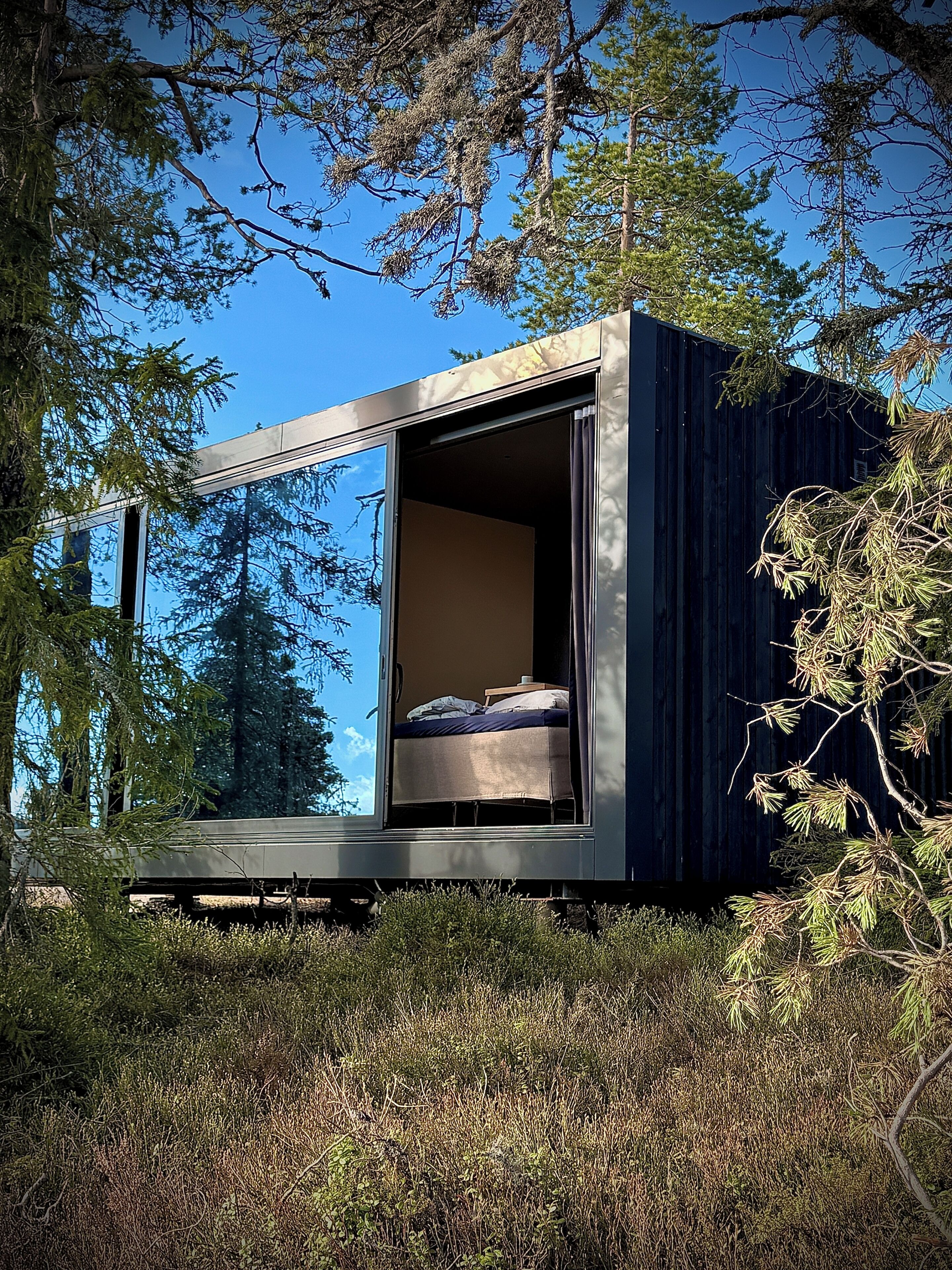 A modern, minimalist cabin with a bed and a small table, surrounded by dense forest.