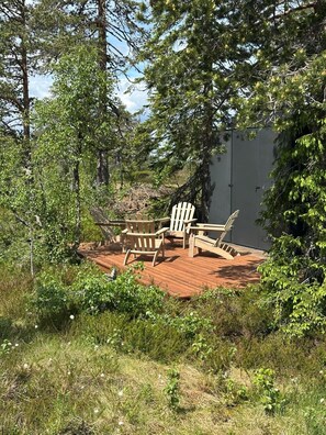 A wooden deck with Adirondack chairs and a bench, surrounded by dense greenery and trees.