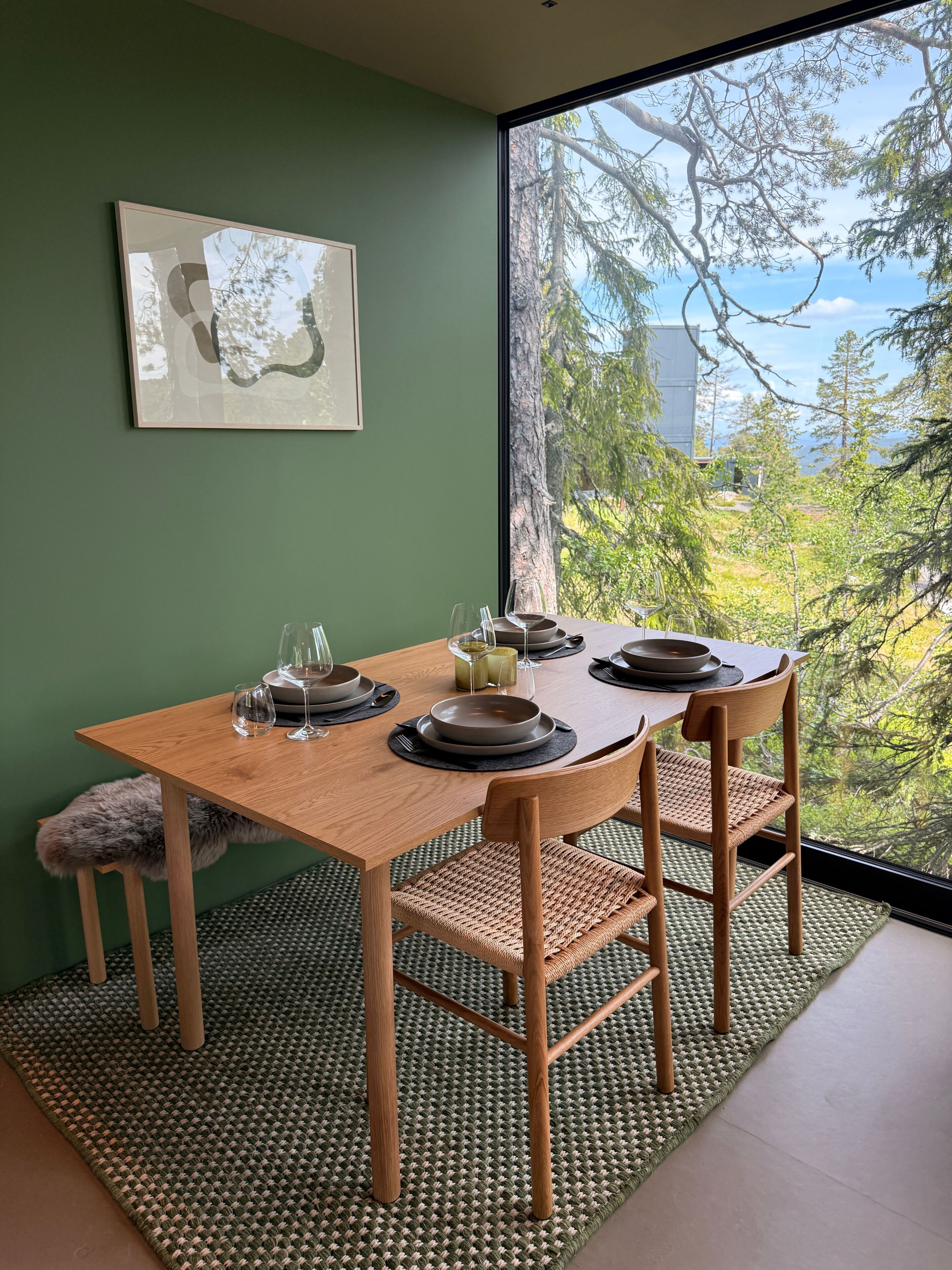 A dining area with a table set for two, a framed picture on the wall, and a view of trees and sky through a large window.