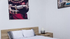 Ein Schlafzimmer mit hölzernem Kopfteil, einem Bett mit weißer Bettwäsche, einem Nachttisch mit Lampe und zwei an der Wand befestigten Kunstwerken, die kulturelle Szenen darstellen.