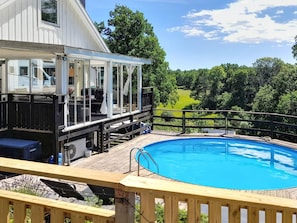 A pool area with a wooden deck, a covered seating area, and a view of a green landscape.