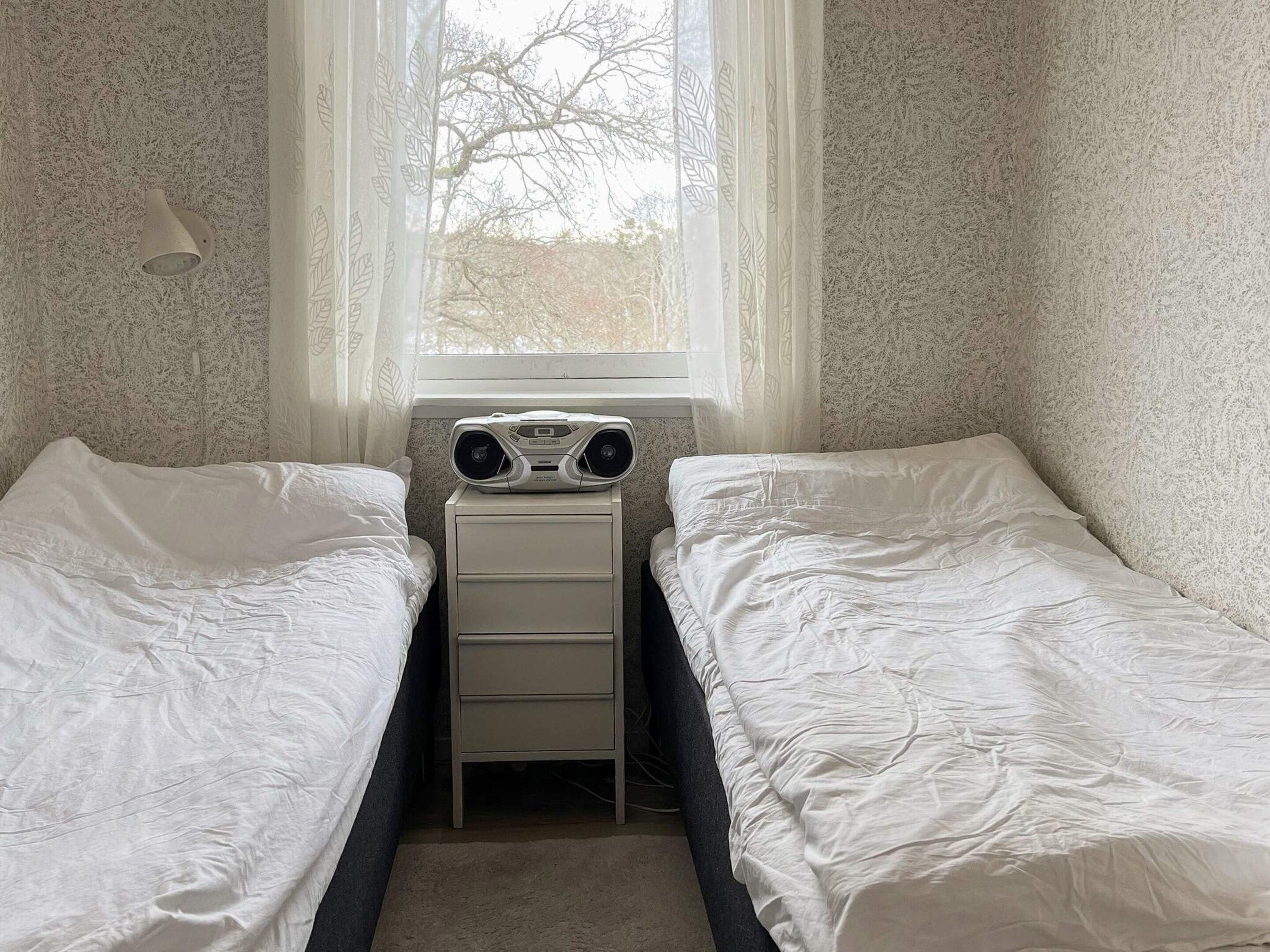 A bedroom with two single beds, a bedside table, a window with sheer curtains, and a wall-mounted lamp.