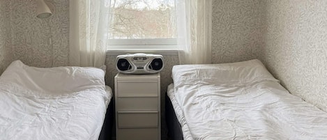 A bedroom with two single beds, a bedside table, a window with sheer curtains, and a wall-mounted lamp.