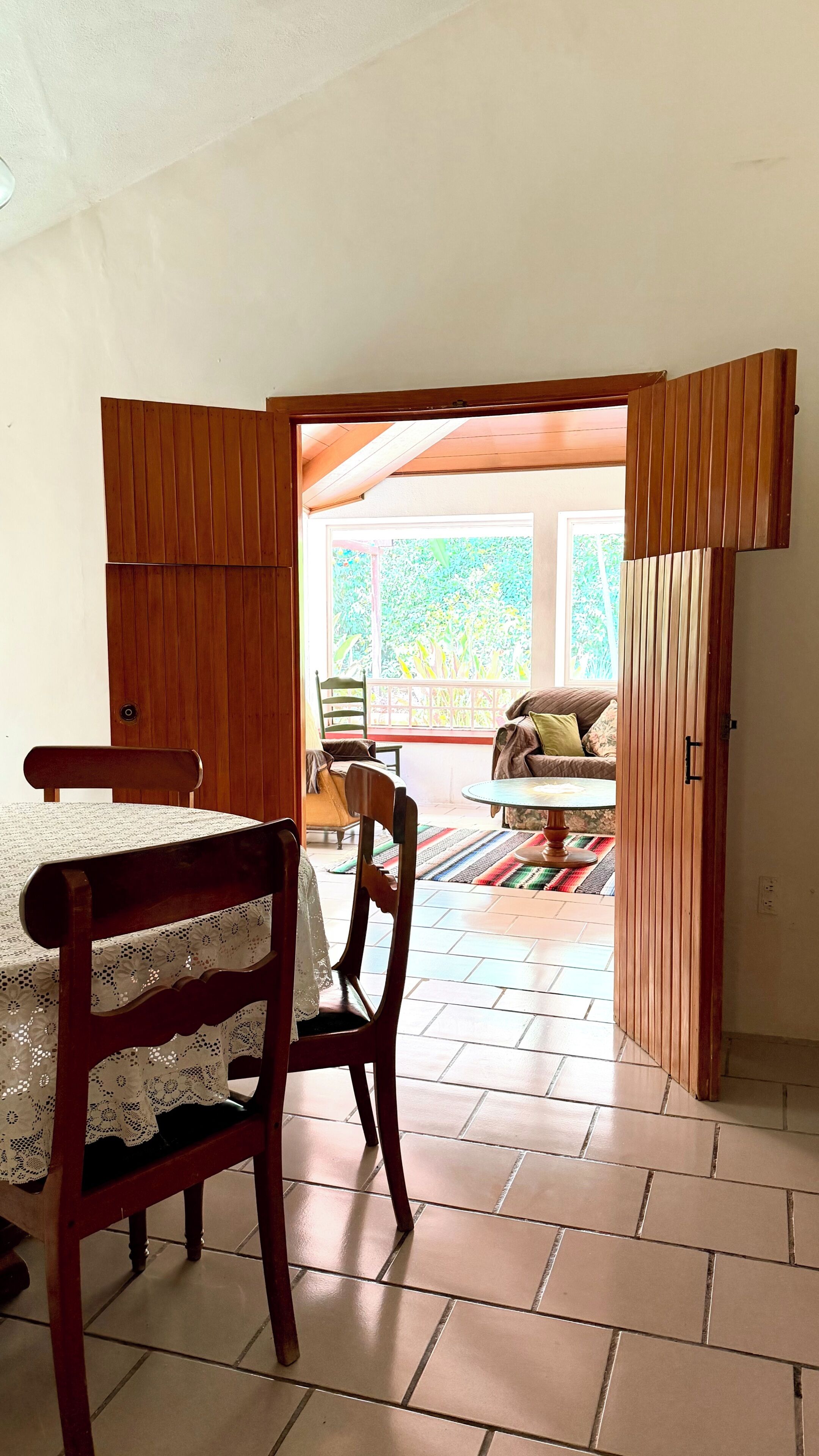 A room with a dining table, chairs, a sofa, and a view through an open door to an outdoor area with a tiled floor and a wooden structure.