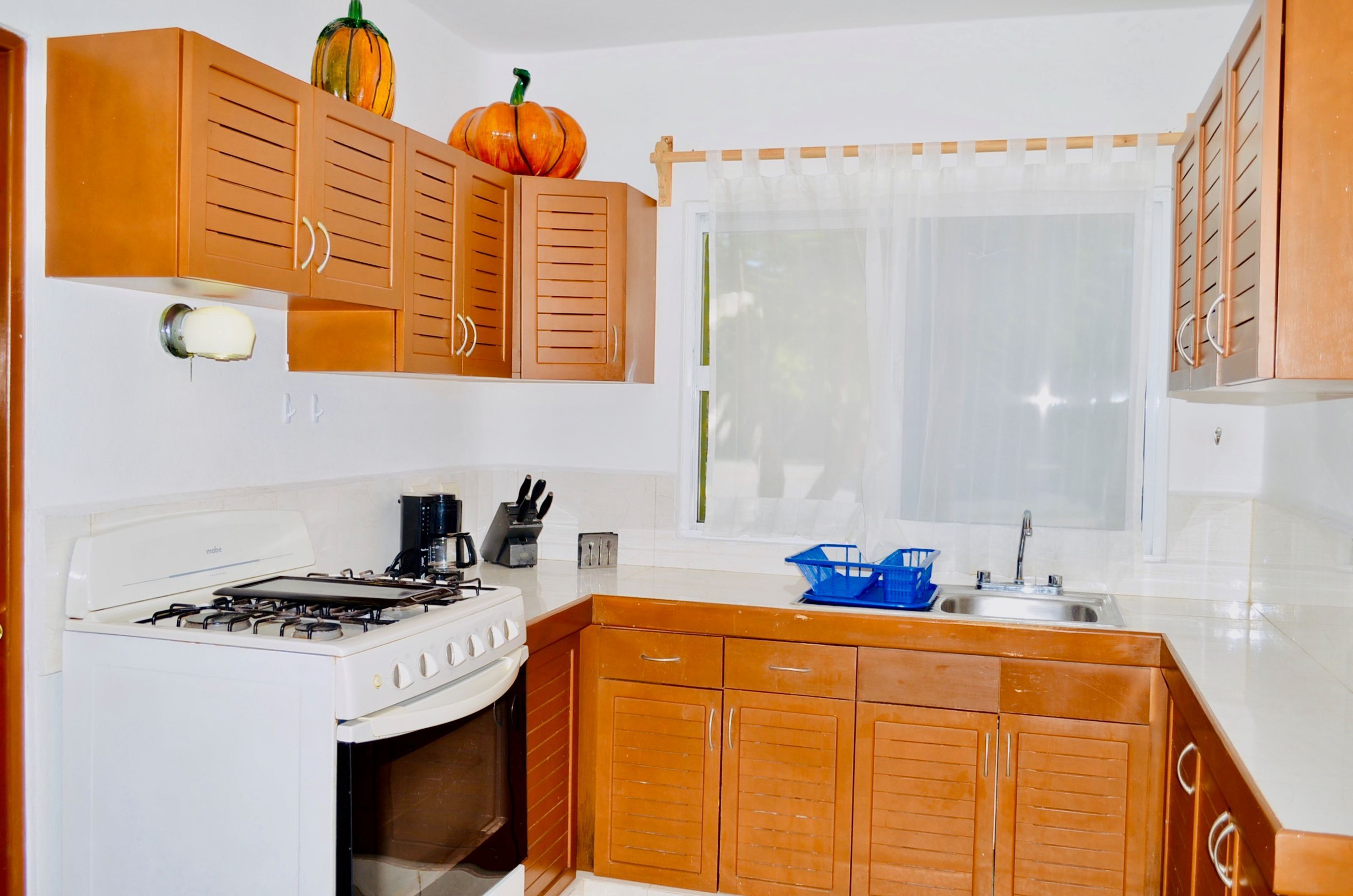 A kitchen with wooden cabinets, a gas stove, a sink, and a window with a sheer curtain.