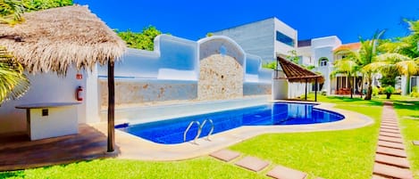 A swimming pool with a thatched roof structure, a stone wall, and a clear blue sky.