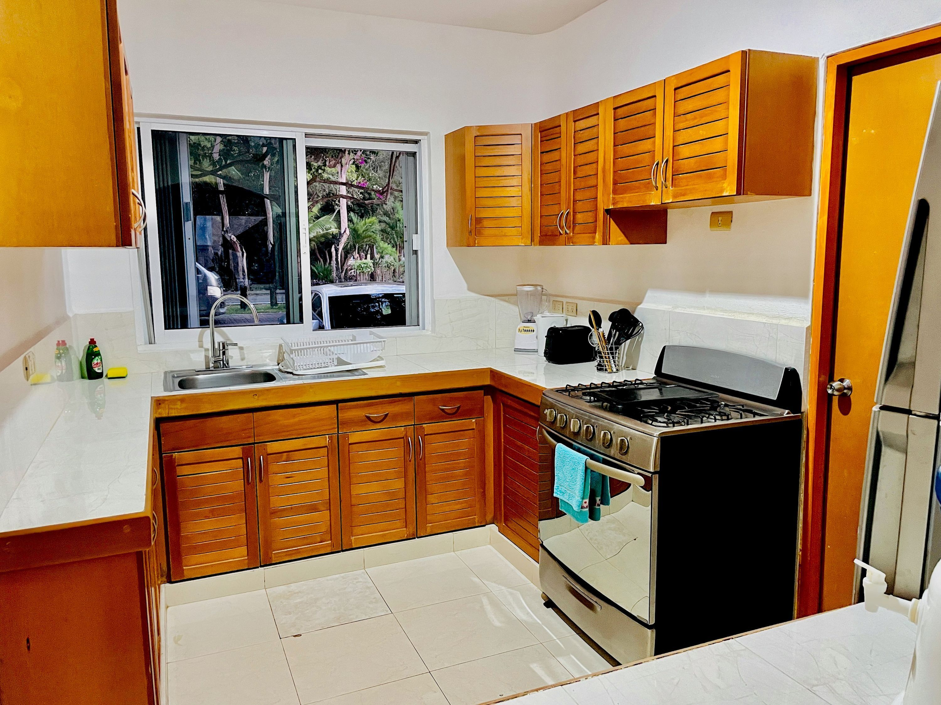 A compact kitchen with wooden cabinets, a gas stove, a refrigerator, and a window with a view of greenery.