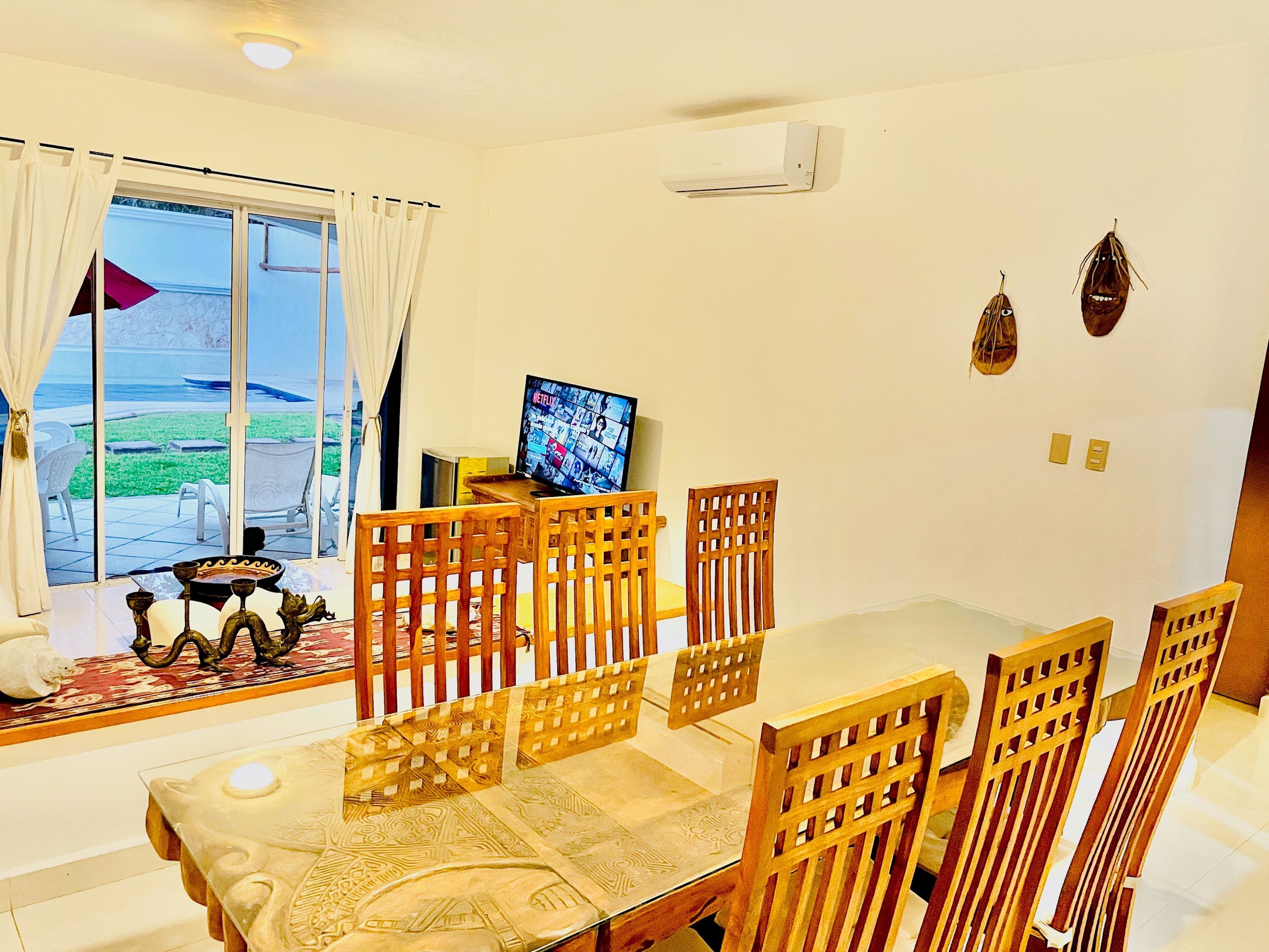 A dining area with a glass table, wooden chairs, a TV, and a view of the outdoors.
