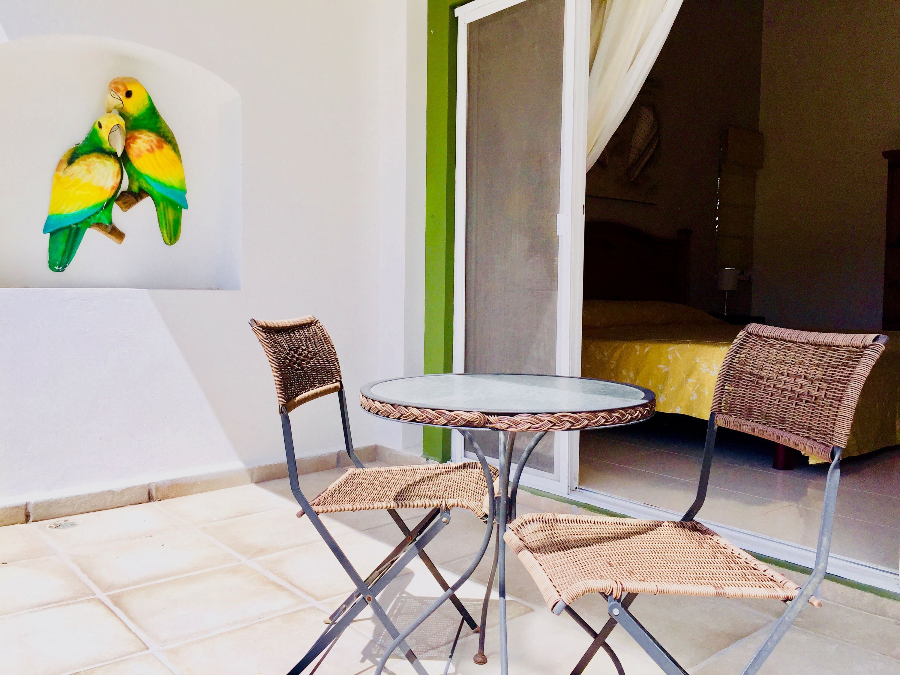 Una habitación con una mesa de vidrio, dos sillas de mimbre, una cama con cubrecama amarillo y un cuadro de pájaros en la pared.