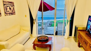 Un salon avec un canapé blanc, une table basse en bois et un téléviseur posé sur un meuble en bois. Une porte-fenêtre coulissante donne accès à une terrasse extérieure avec vue sur la mer.