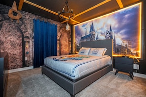 Ein Schlafzimmer mit Backsteinwand, einem großen Poster zum Thema Hogwarts, einem Bett mit weißblauer Bettdecke und einem schwarzen Nachttisch mit Lampe.
