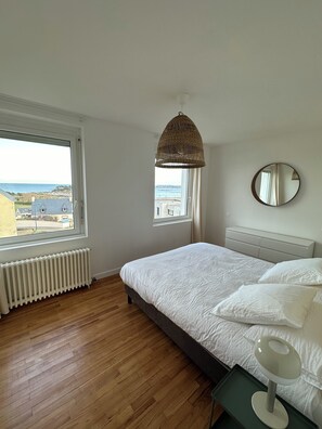A bedroom with a large window, a bed, a bedside table, a lamp, a mirror, and a ceiling light. - Les Bords de Mer, Saint Pol de Léon (Saint-Pol-de-Léon)