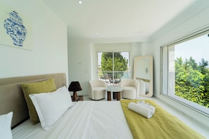 A modern hotel room with a large bed, two armchairs, a small table, a mirror, and a view of greenery through the window. - Villa Punique: Villa exceptionnelle avec vue mer (Site archéologique de Carthage)