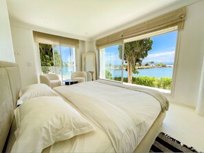 Une chambre avec un grand lit, un coin salon avec des fauteuils et une vue sur un plan d’eau.