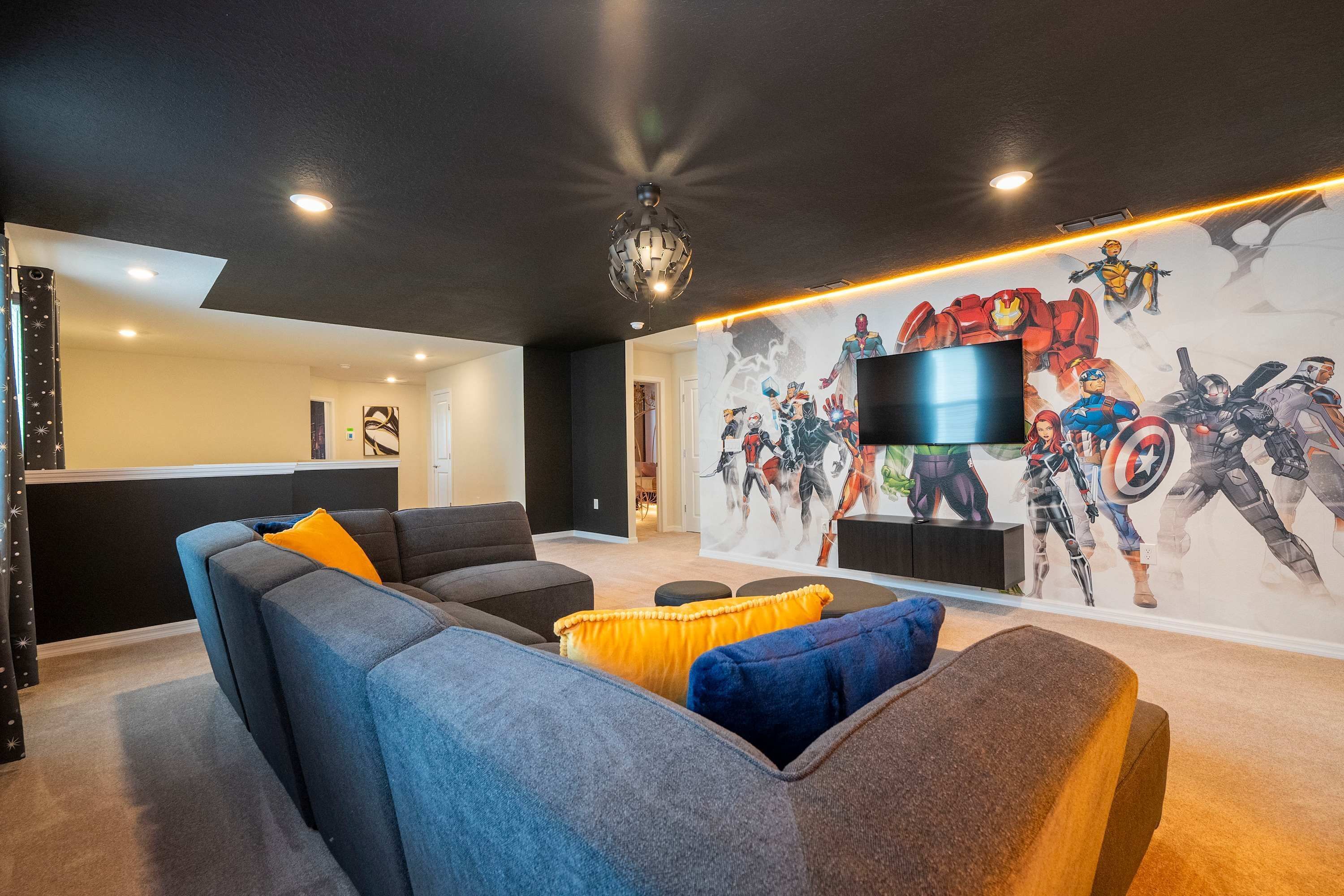 Un salon moderne avec un grand canapé d’angle, un téléviseur à écran plat fixé au mur et une fresque représentant des super-héros.