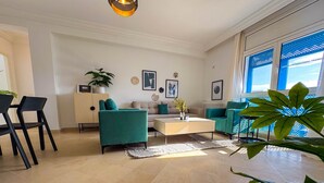 Ein modernes Wohnzimmer mit einer Couch, einem Couchtisch und Sesseln. Eine Pflanze, eine Kommode und Kunstwerke sind an den Wänden zu sehen.