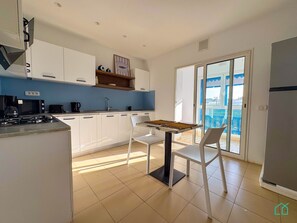 A modern kitchen with white cabinets, a gas stove, a microwave, and a coffee maker. There is a dining table with two chairs and a view of a balcony with a blue railing. - Coste Sidi Bou Said A3: Spacious and central (Site archéologique de Carthage)