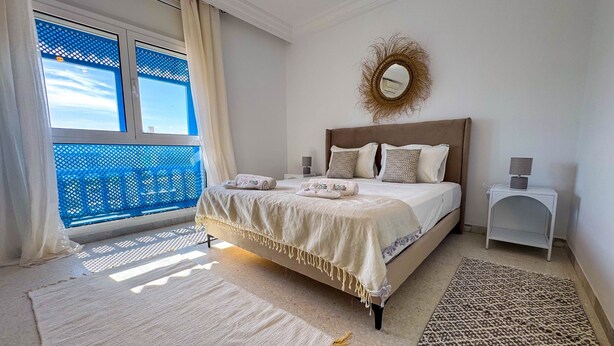 Ein Schlafzimmer mit Bett, Nachttisch, Spiegel und Blick zum Himmel durch ein Fenster.