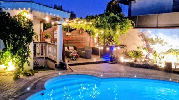 Ein Schwimmbecken mit Steinbord, einer Holzterrasse und einer Pergola mit Lichterketten.