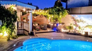 Une piscine bordée de pierres, une terrasse en bois et une pergola illuminée de guirlandes lumineuses.