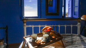 Una habitación con una cama, un ventanal con contraventanas azules y una bandeja con tazas y un bol sobre la cama.
