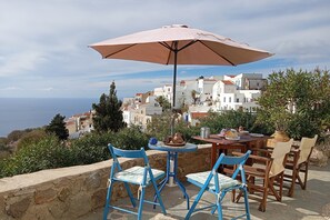 A terrace with a table and chairs, a large umbrella, and a view of white buildings and the sea.