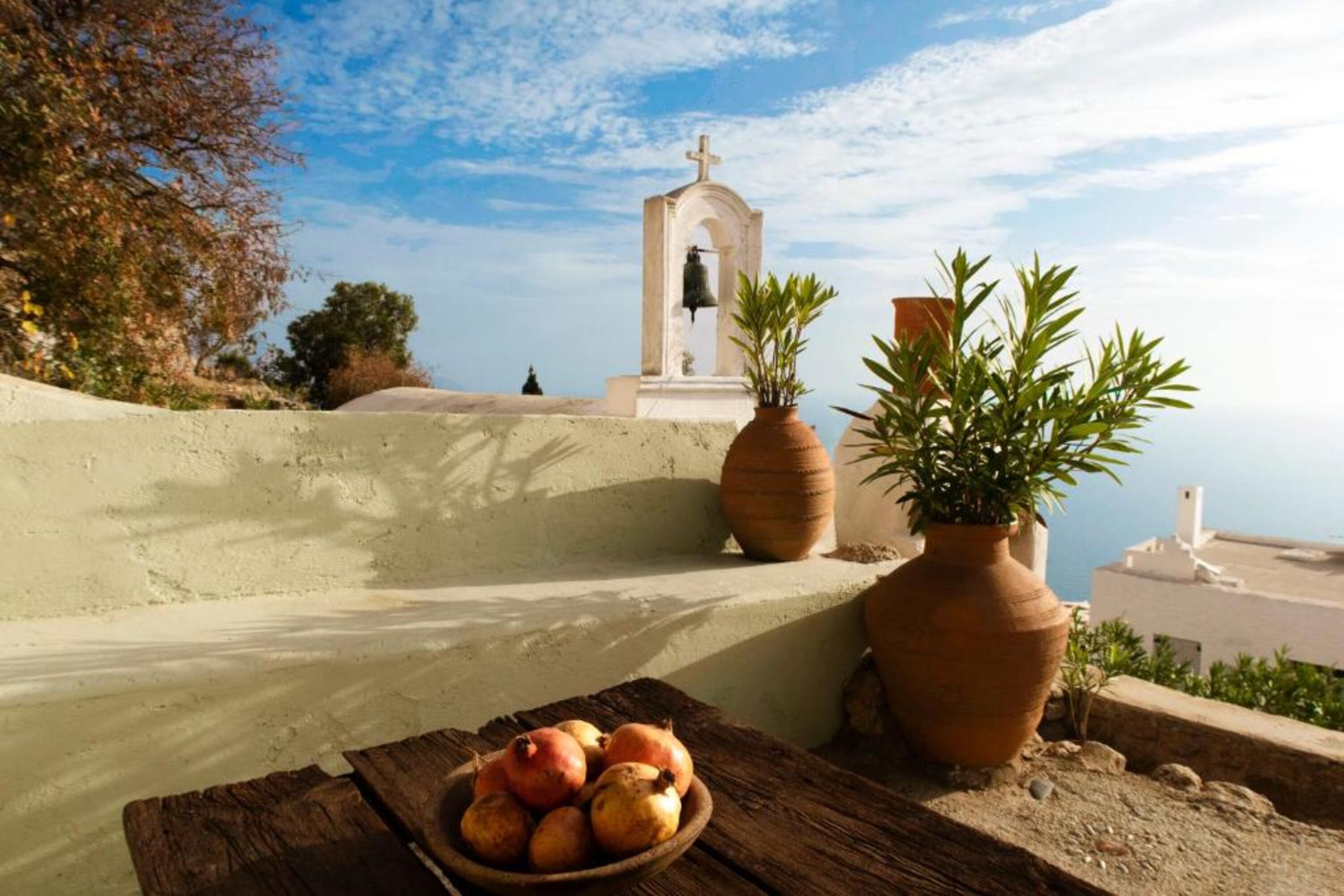 Une vue sur la côte, avec un clocher, des plantes en pot et une table en bois sur laquelle se trouvent des fruits.