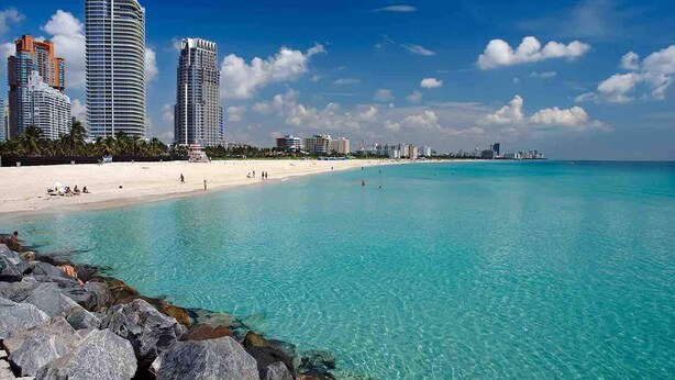 Een strand met helder turquoise water, een zandstrand en hoge gebouwen op de achtergrond.