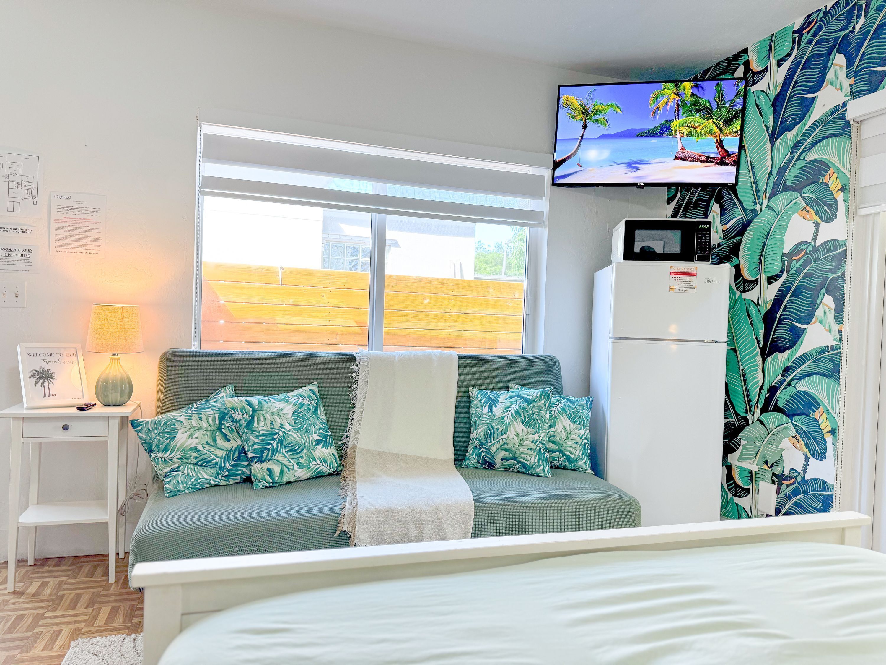 A room with a bed, a TV mounted on the wall, a window with a view of a wooden fence, and a refrigerator.