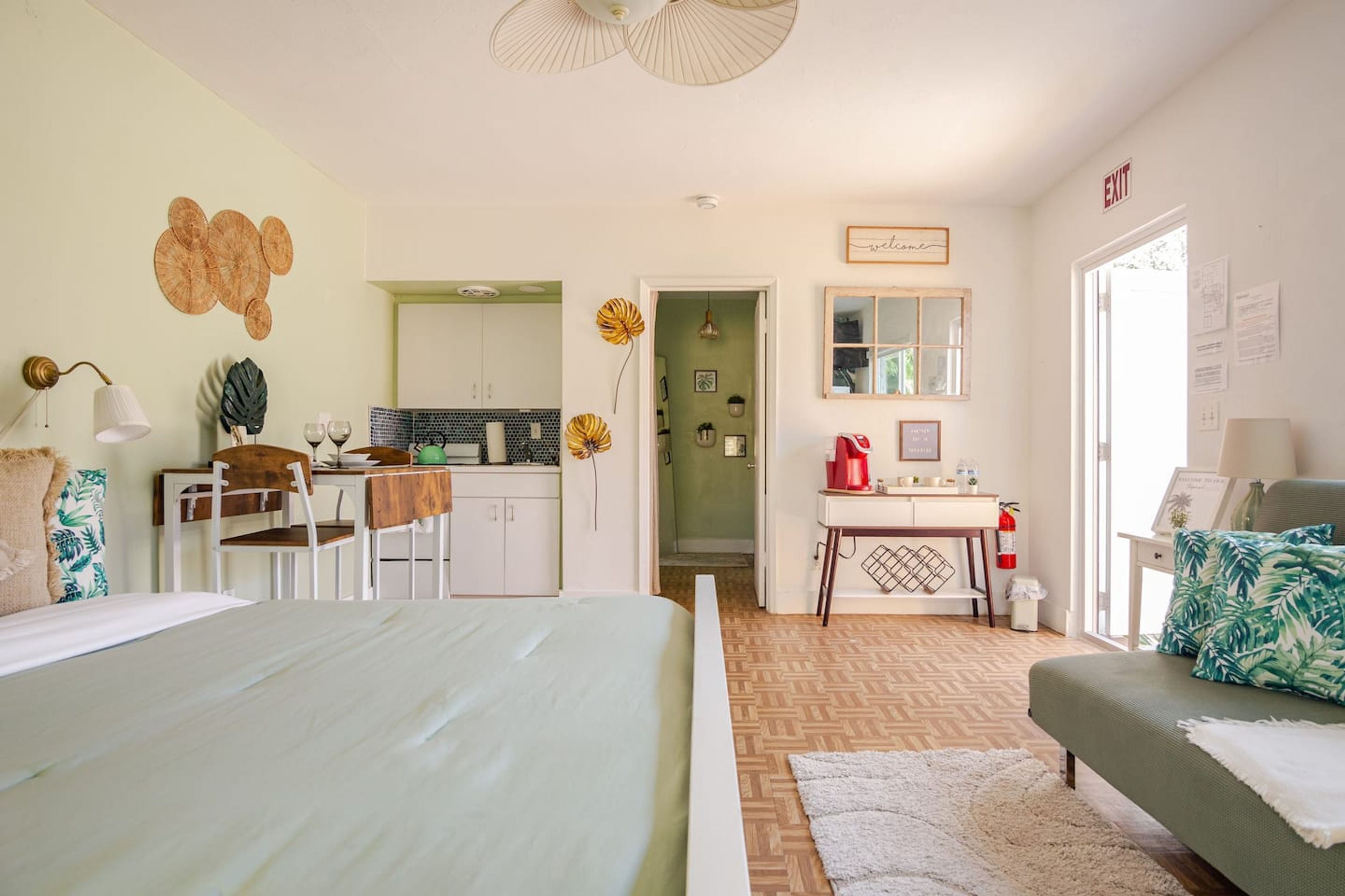 A bedroom with a bed, a sofa, a desk, and a kitchenette in the background.