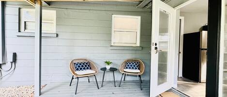 A covered patio area with two wicker chairs and a small table, a door leading inside, and a concrete floor.