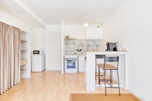Une cuisine compacte avec des meubles blancs, un four à micro-ondes, une cuisinière et une machine à café. On y trouve deux tabourets de bar et un petit îlot central avec un évier.