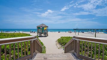A wooden staircase leading to a beach area with a lifeguard tower, beach umbrellas, and a pier extending into the water.