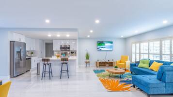 A modern kitchen with stainless steel appliances, a dining area with a blue sofa and yellow armchairs, and a TV mounted on the wall.