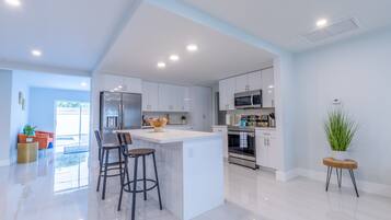 A modern kitchen with white cabinets, a central island with stools, stainless steel appliances, and a large window providing natural light.