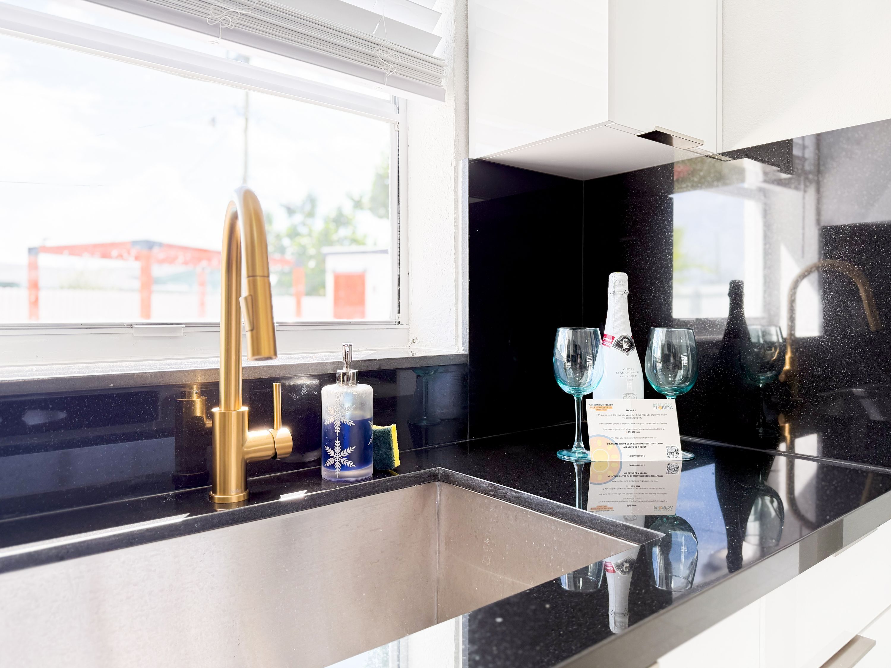 Een moderne keuken met een zwart aanrecht, een roestvrijstalen spoelbak, een gouden kraan en een raam met uitzicht op een rode constructie.