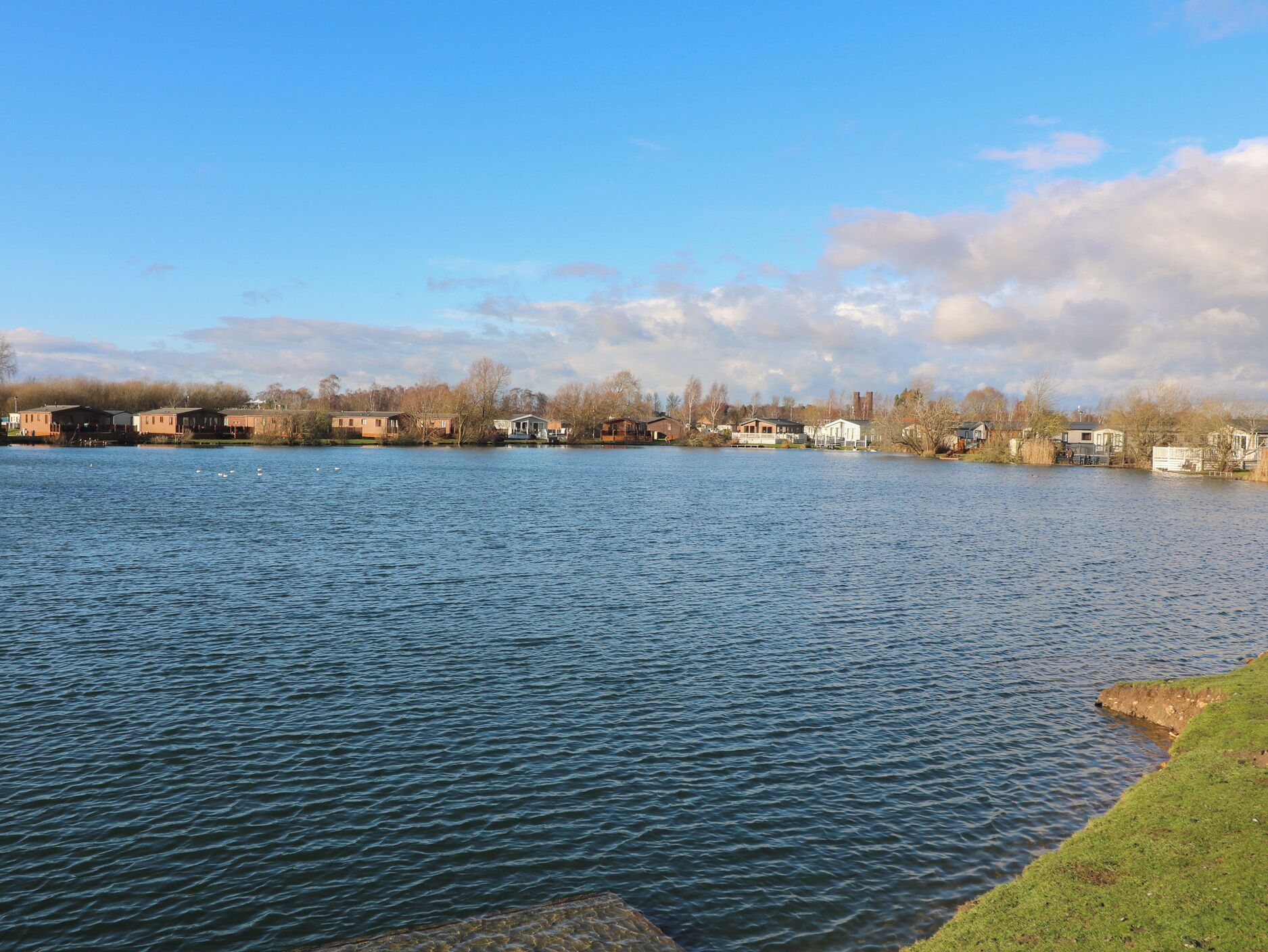 A calm lake with houses along the shore, a clear sky, and some trees in the background.