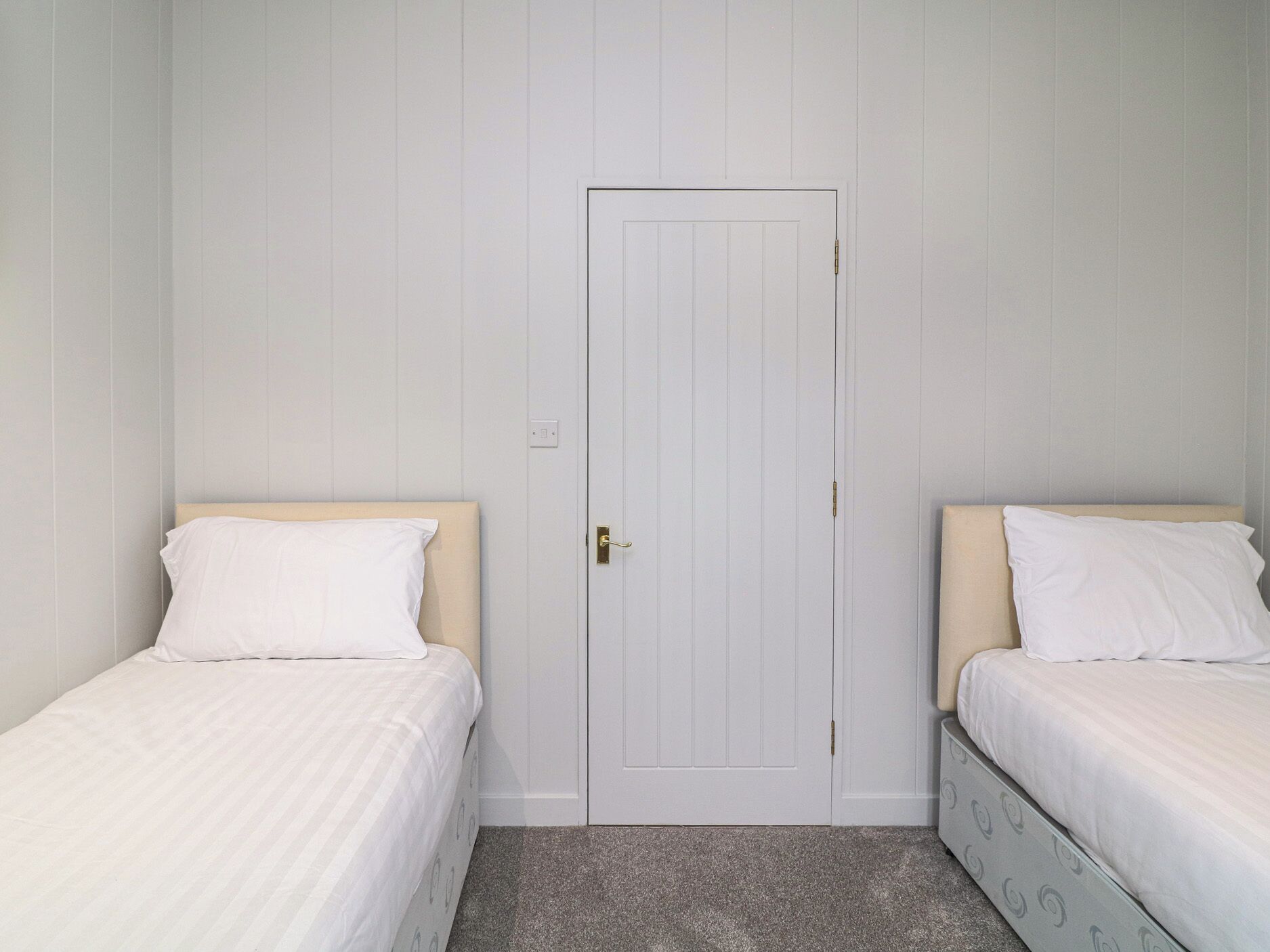 Une chambre avec deux lits simples, une porte blanche et un sol recouvert de moquette.