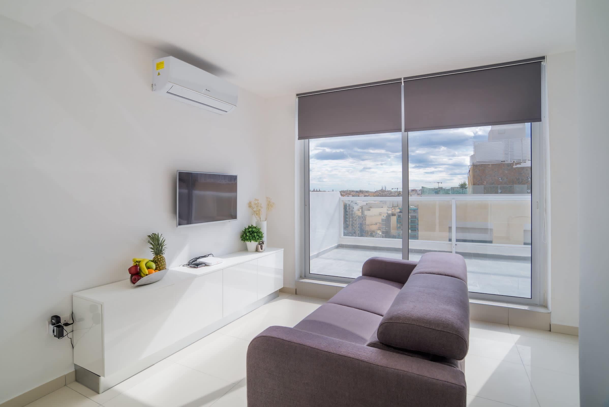 A modern living room with a grey sofa, a flat-screen TV, a white cabinet, and a large window with a city view.