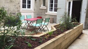 A courtyard with a red table and green chairs, surrounded by plants and a wooden planter.