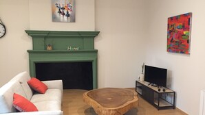 A living room with a green fireplace, a white sofa with red cushions, a wooden coffee table, a TV on a black stand, and a colorful abstract painting on the wall.