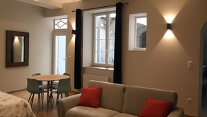A modern living room with a beige sofa, two red cushions, a dining table with chairs, and a large window with curtains.