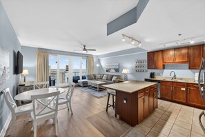 Eine moderne Küche mit Holzschränken, ein Essbereich mit weißem Tisch und Stühlen sowie ein Wohnzimmer mit Sofa und Couchtisch.