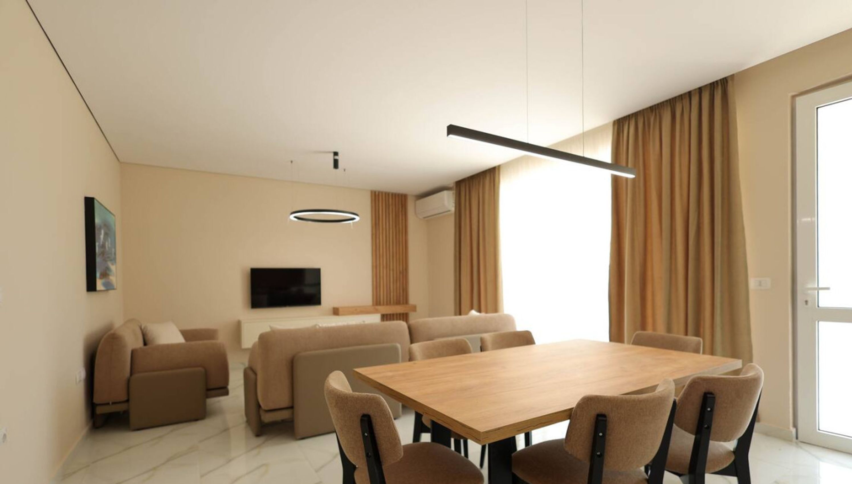 A modern living room with a wooden dining table, beige sofas, and a flat-screen TV mounted on the wall.