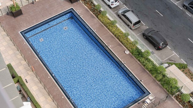 Una piscina rectangular con un diseño de mosaicos azules, rodeada por un camino pavimentado y bordeada de follaje.