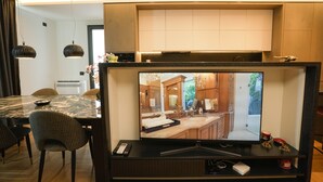 En moderne stue med et stort fladskærms-tv monteret på en trævæg, et spiseområde med et marmorbord og mønstrede stole, og et køkken, der kan ses gennem en åben dør.