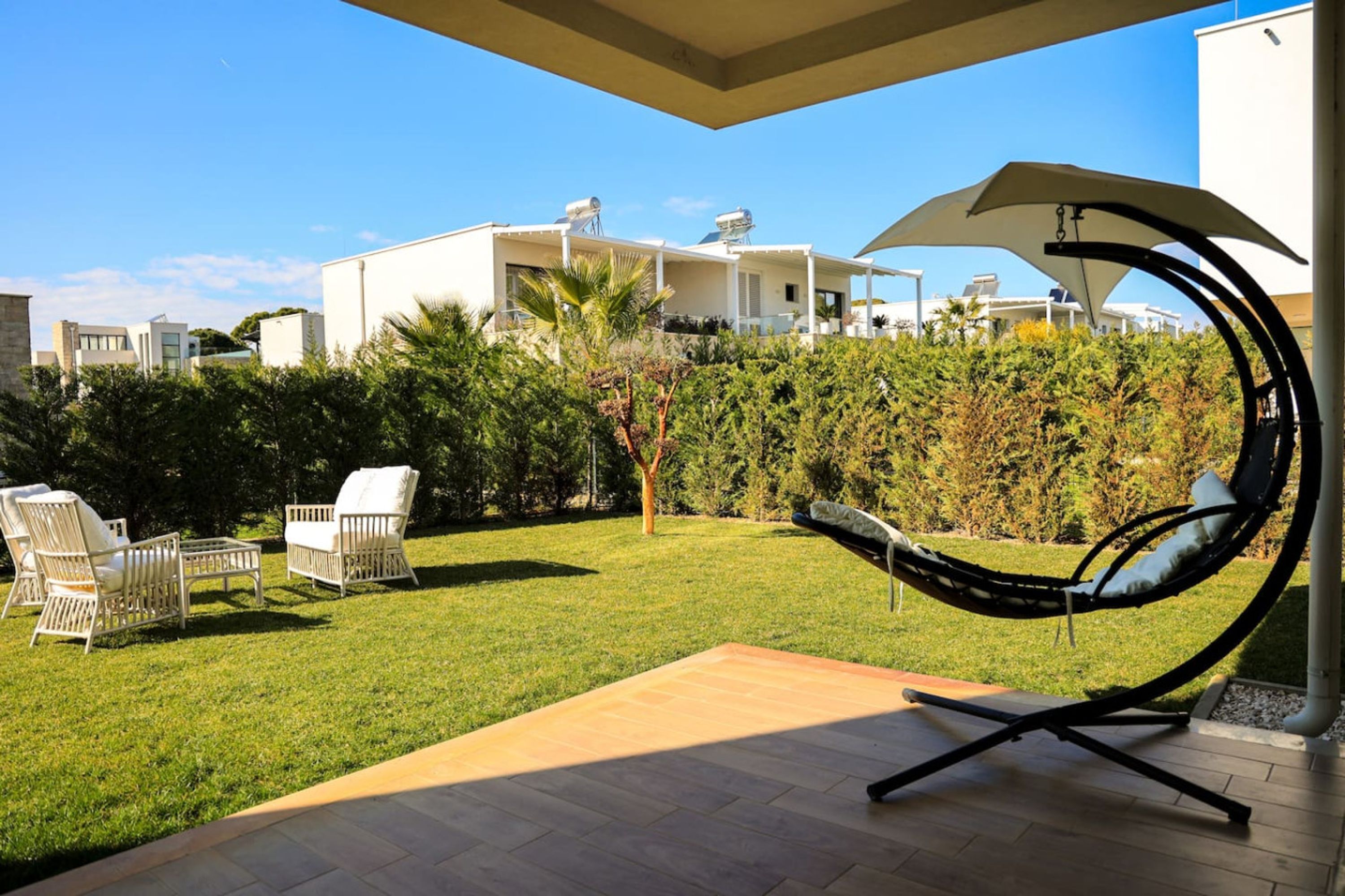 A modern outdoor patio with a swing chair, white wicker furniture, and a view of a white building in the background.