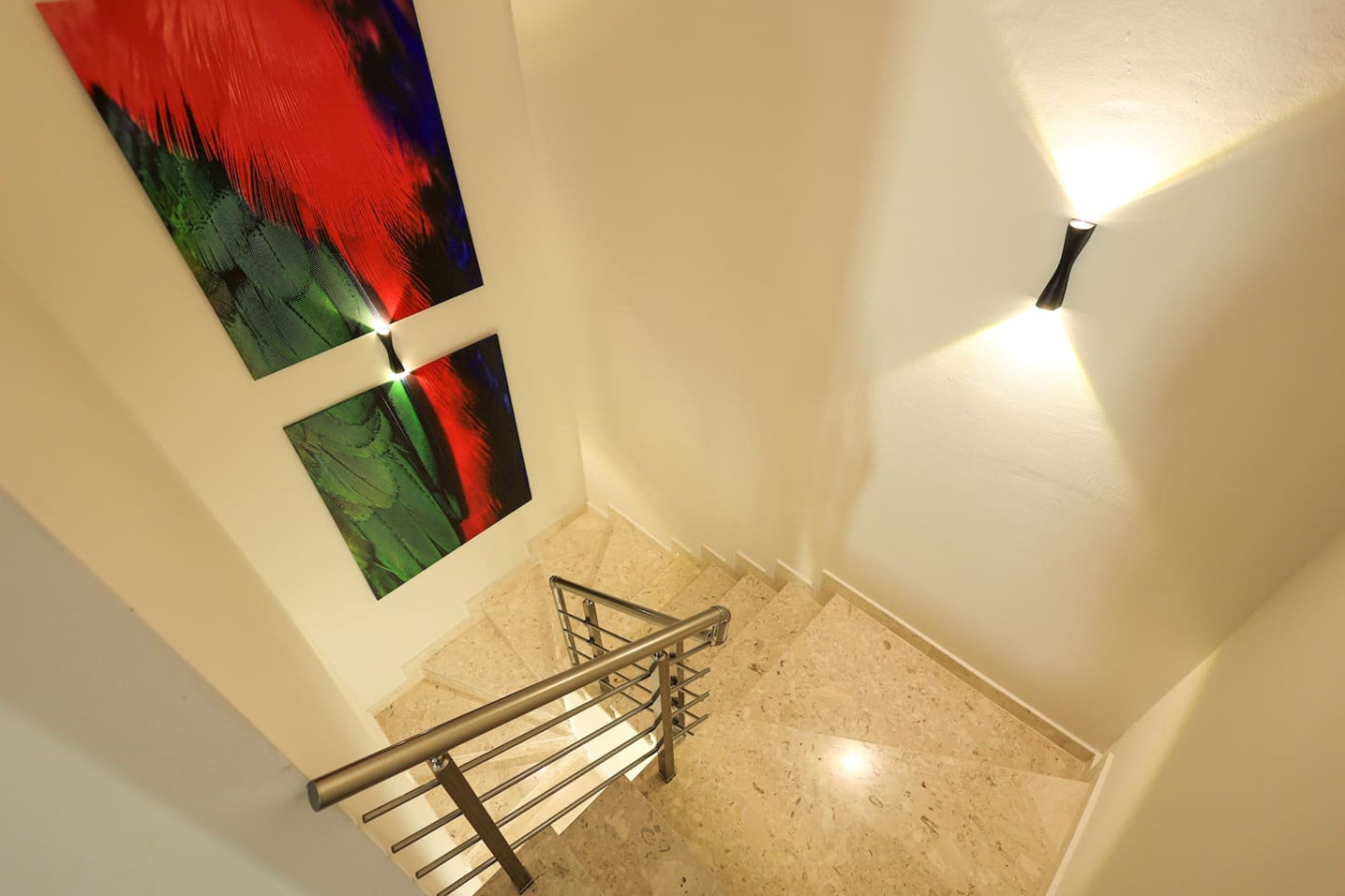 Una escalera con barandilla metálica, un accesorio de iluminación en la pared y dos cuadros abstractos en la pared.